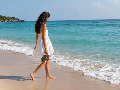 A woman in a white dress walking barefoot along a beach